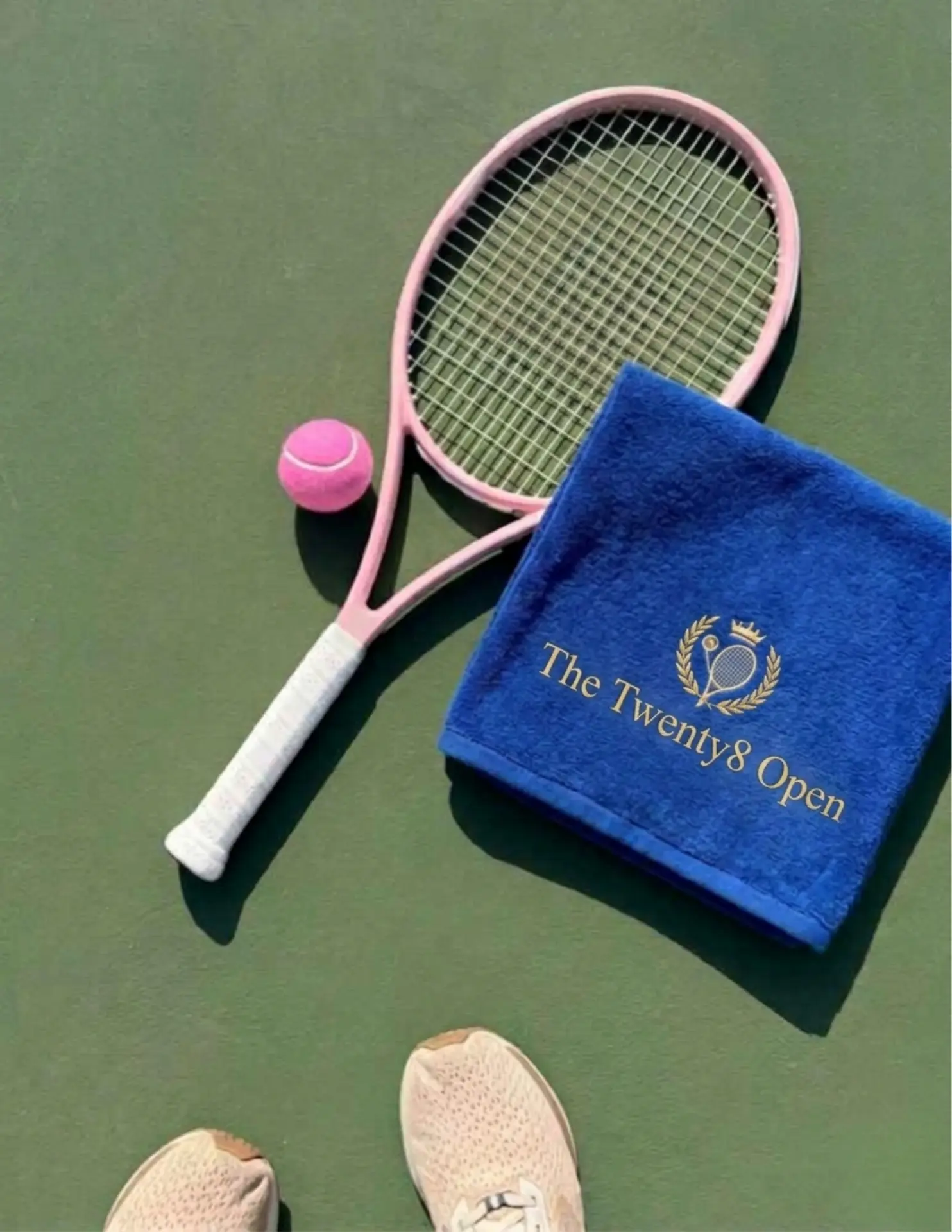 Top-down pink tennis racket, pink ball, navy Twenty8 Open towel and athletic shoes on a green hard court