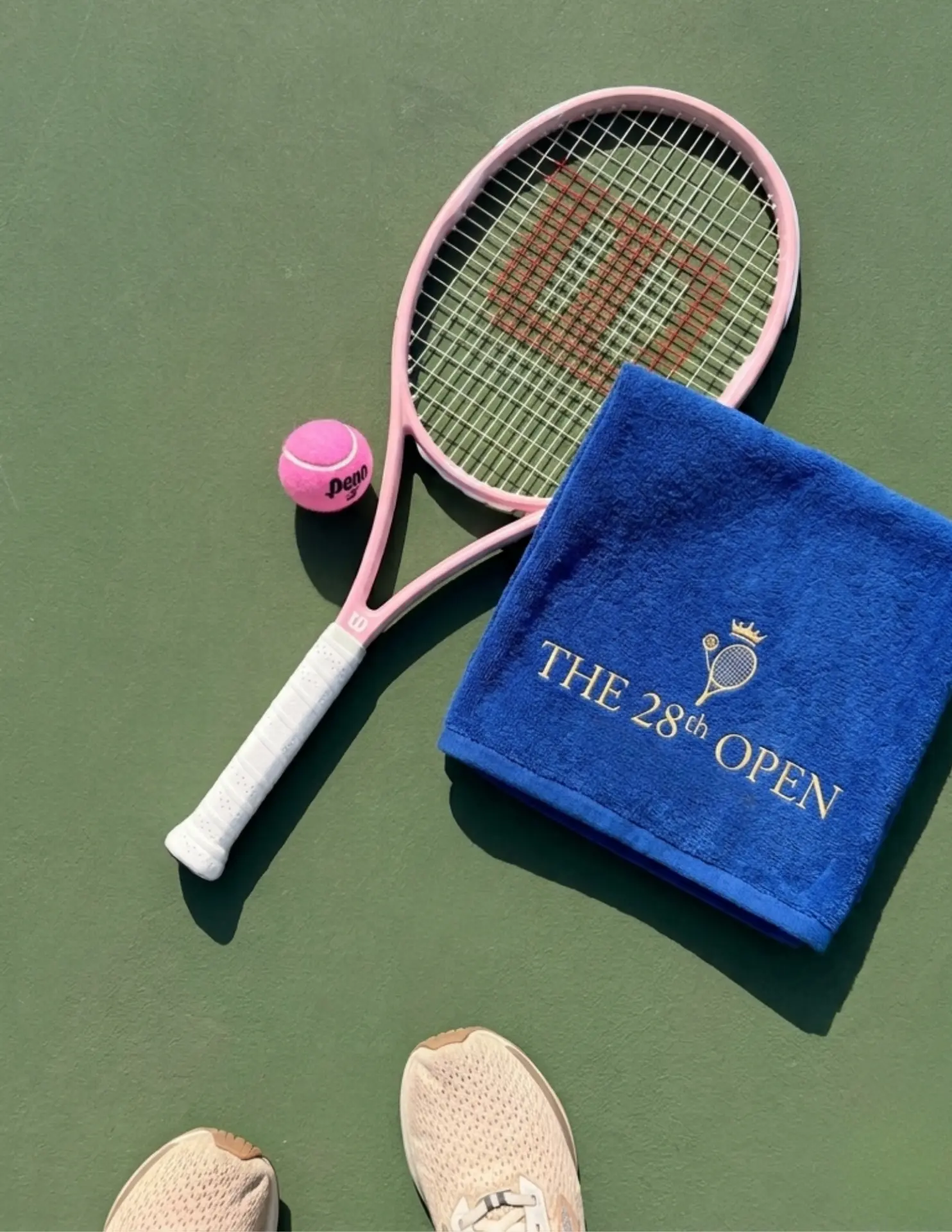 Top-down pink tennis racket, pink ball, navy 28th Open towel and athletic shoes on a green hard court